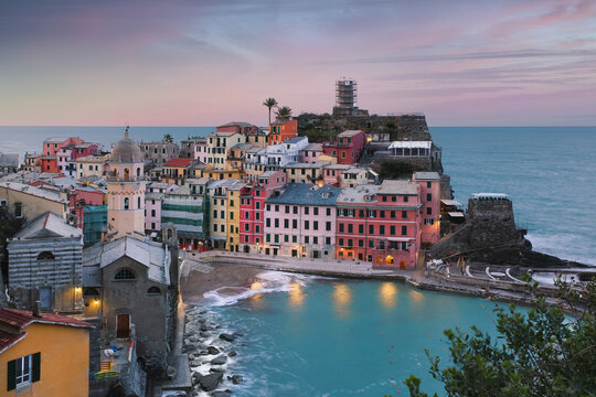 View of the colorful houses and the historic church of Santa Margherita di Antiochia at twilight in Vernazza, Liguria, Italy.