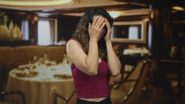 Young hispanic woman anxiously covers face while seated at polished wooden table in elegant restaurant dining area; stress.