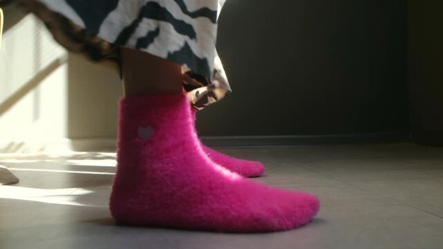 Low angle view of someone feet wearing cozy, furry pink socks with love symbol and zebra print pajama pants, dancing on floor at home, illuminated by warm morning sunlight coming from window