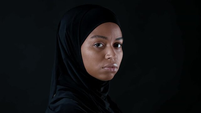 A young woman with dark skin wears a black hijab. Her expression is serious as she gazes directly at the camera. The background is dark, enhancing her presence.