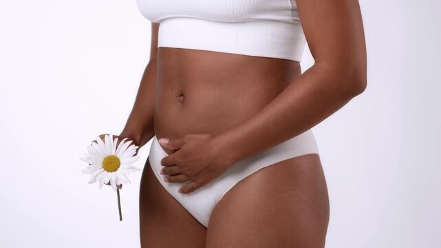 A woman with dark skin stands confidently while holding a daisy flower. She is wearing simple white clothing and poses in a neutral space, showcasing a natural look and body positivity.