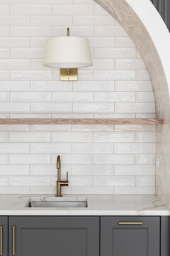 A modern kitchen sink detail with a brass faucet against a white subway tile backsplash, floating natural wood shelf, dark gray cabinets,, gold light fixture, and pattern wallpaper.
