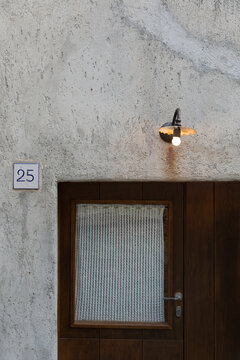 View of a wooden door with a lace curtain and a vintage lamp on a rough white wall in Salina, Sicily, Italy.