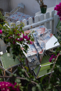 View of a small outdoor table with a nautical-themed tablecloth and green chairs behind a white picket fence and pink bougainvillea flowers in Salina, Sicily, Italy.