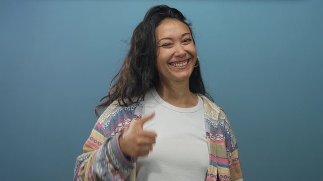Woman pointing finger and giving thumbs up to camera with visible hand and smile in blue studio; confidence encouragement.