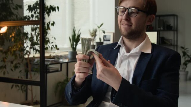 Tilt up view of excited formally dressed young man taking stack of money from white table, bringing it close to his face and kissing with delighted expression during workday in office setting