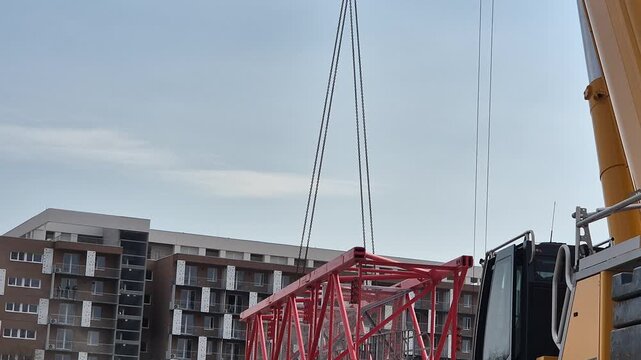 Rear view of a mobile crane lifting a tower crane mast section, showing the operator's cabin and the initial separation from the ground.