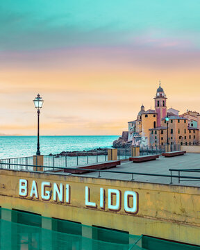 View of the Bagni Lido terrace and the Basilica di Santa Maria Assunta against a sunset sky in Camogli, Liguria, Italy.