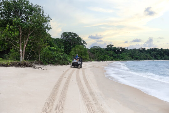 Gabon - 09 April 2023: View of an off-road vehicle driving along a sandy beach with tourists, lush tropical forest, and ocean waves under a cloudy sky.