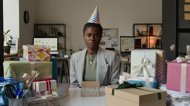 Zoom out portrait of indifferent Black young woman with birthday hat posing for camera at office table cluttered with wrapped gifts