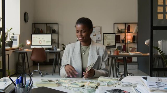 Zoom in shot of African American girl excitedly examining stacks of dollar bills and smelling them while sitting at messy work table in office setting