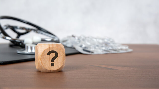 Question mark on wooden block with blurred stethoscope and pills. Medical advice, health diagnostics and healthcare consultation concept.
