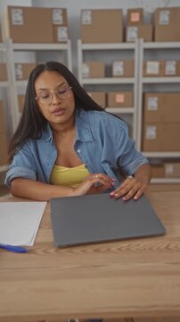 Woman with glasses in denim shirt closes laptop with hand and rubs forehead at a storage building with shelves of boxes; small business fatigue.