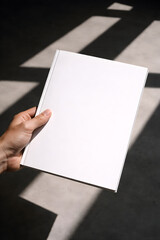 Naklejka premium Hand holds blank book against sunlit concrete floor