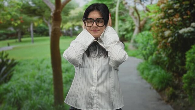 Woman wearing white shirt and glasses covers ears amid lush green forest under dappled sunlight at dawn; irritation.