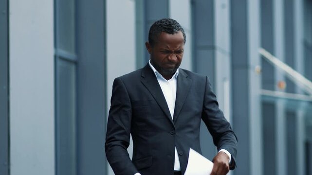 A man with dark skin is standing outside an office building. He is wearing a black suit and reading through several papers. The setting is urban, with modern architecture in the background.