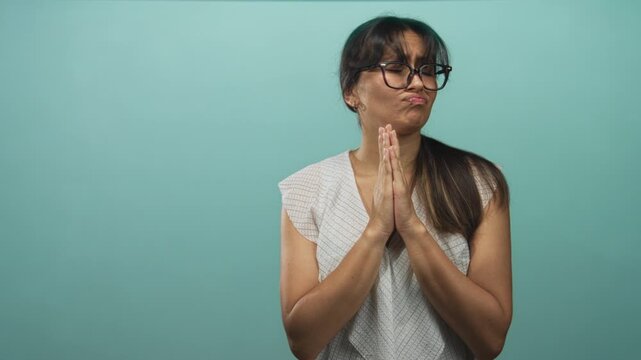 Hispanic young brunette woman pressing hands together in teal studio, wearing glasses and a white blouse; pleading forgiveness.