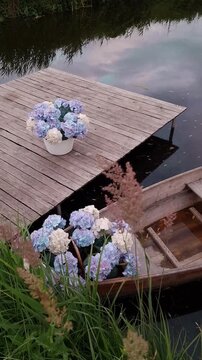 Old wooden rowing boat floating on dark water near a pier, decorated with white and blue hydrangea bouquets, evening light, reflection on water, romantic atmosphere.