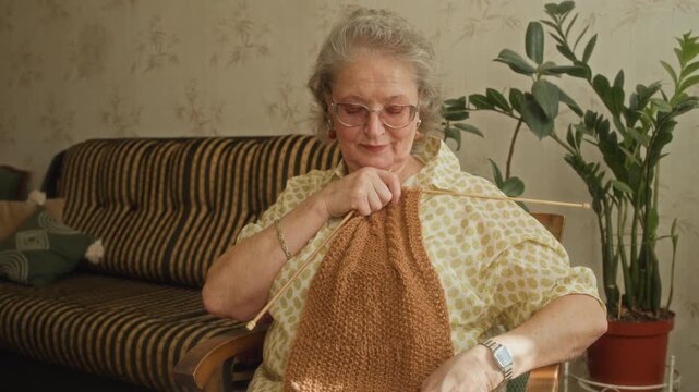 Medium shot of smiling elderly woman enjoying with white hair trying on handmade wool scarf while knitting with needles sitting in cozy armchair in retro sunlit living room