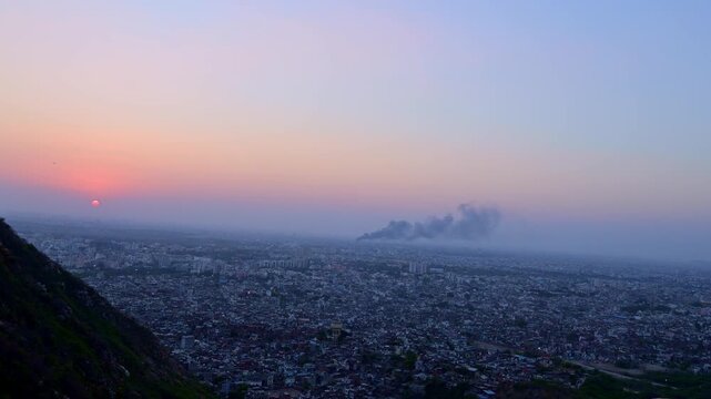 Sunset view from Nahargarh Fort Jaipur showing orange sun near horizon, Jaipur skyline, evening sky with pink-blue gradient, buildings and smoke in distance.