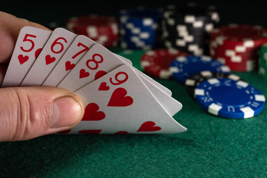 A man holds playing cards with hearts or a straight flush in his hand. In the background are stacked poker chips. The room is well lit, the game is lively.
