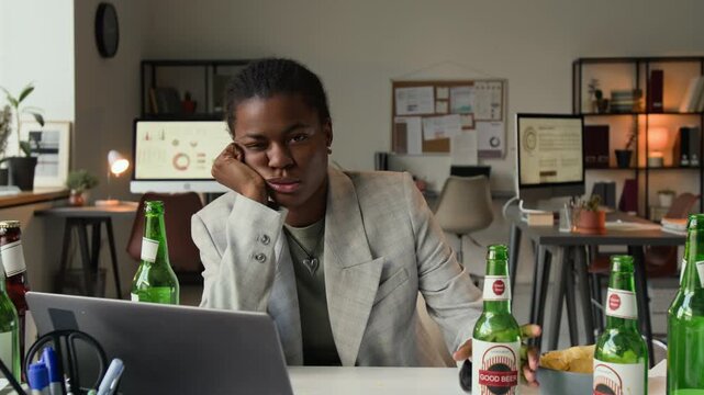 Zoom in portrait of fatigued African American girl in grey jacket drinking beer and looking at camera indifferently during workday in office