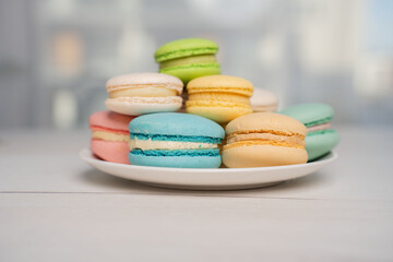 Obraz premium close-up of a pastel assortment of colorful french macarons stacked on a white plate on a pale wooden table with soft natural bokeh background, creating a delicate airy dessert scene, shallow