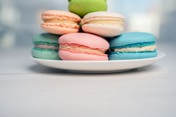 Obraz premium close-up of pastel macarons stacked on a white plate on a pale wooden tabletop, soft airy high-key composition showcasing pink, blue, green and cream cookies with delicate textured fillings