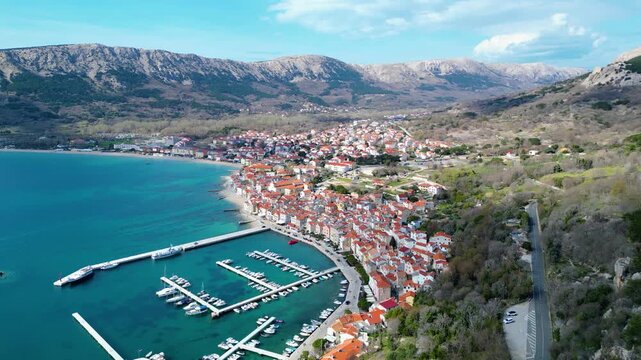 Aerial view of Baska's vibrant harbor and red-roofed town nestled against dramatic mountains on Krk Island, Croatia. Clear skies and turquoise waters.