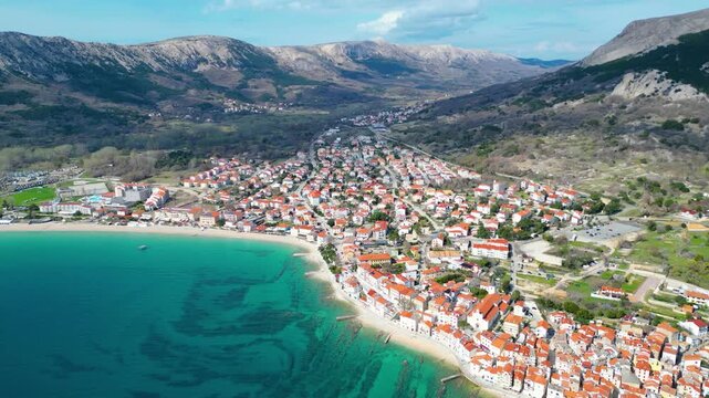 Aerial view of Baska, Krk Island, Croatia, showcasing its vibrant red-roofed town, turquoise Adriatic Sea, and dramatic mountains under a clear spring sky.