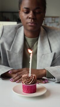 Vertical view of apathetic African American office girl blowing out single candle on birthday cupcake sitting at work desk