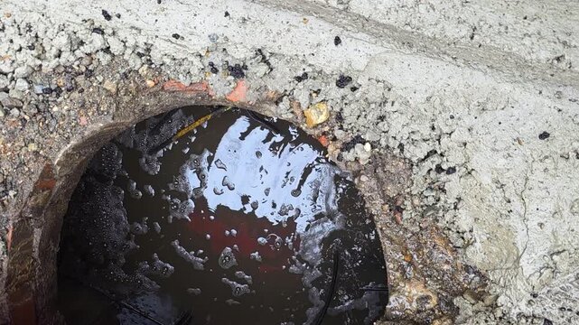 Murky Concrete Drain Opening With Water Oil Sheen And Leaf Litter Cling To Gritty Sediment Around Circular Lip, Reflective Surface Shows Overcast Sky, Sidewalk Outlet Detail, Atmospheric