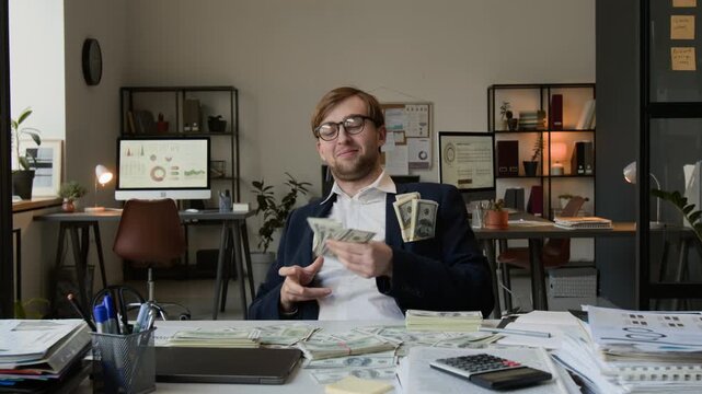 Portrait of money driven young man wearing glasses and suit smugly throwing stacks of banknotes on work table looking at camera, taking them from pockets of his jacket