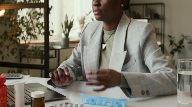 Tilt up view of busy Black business girl fumbling for blue pill organizer on white desk without looking at it and taking medicine while working on laptop
