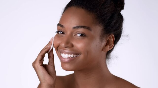 Woman with dark skin is using a cotton pad to apply a skincare product on her face. She is smiling and looks happy in a bright, well-lit setting.