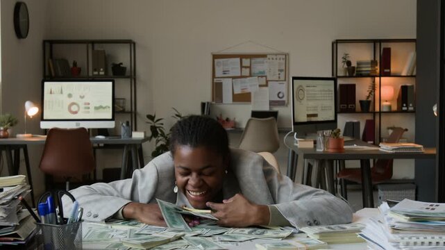 Zoom in view of overjoyed African American gen Z woman resting her chin on office desk ecstatically grabbing dollar banknotes