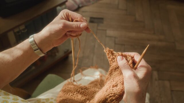 Over shoulder shot of unrecognizable elderly woman unravelling wool scarf pulling out yarn to fix mistake while knitting handcrafted garment in sunlit living room