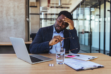 Fototapeta premium Tired Businessman At Office Desk Rubbing Forehead With Glasses Laptop And Documents