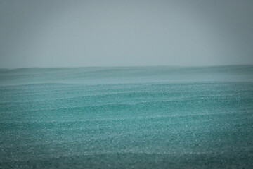 CLOSE UP: Rain and sea mist drift across a calm ocean surface under a pale gray sky. Minimal coastal scene with layered water texture, distant horizon and moody tropical weather on Indonesian island. © helivideo