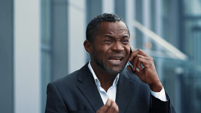 A Black man in a suit is talking on his phone outside a modern building. He appears engaged in a conversation, using hand gestures to emphasize his points.