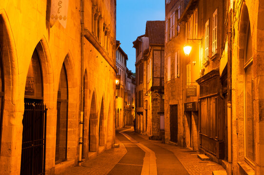 Explore the narrow streets of Figeac at dusk with warm lights illuminating the historic architecture of Emile Zola Street