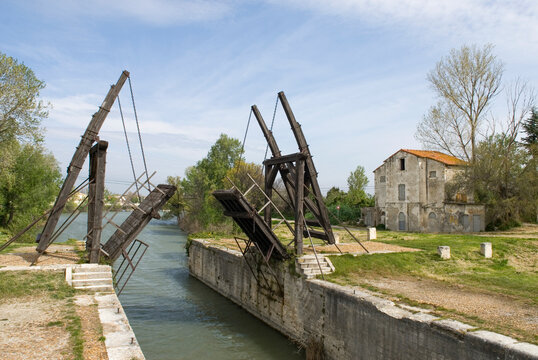 Langlois Bridge in Arles shows its iron structure over a waterway surrounded by green trees and a historic building in the background, France
