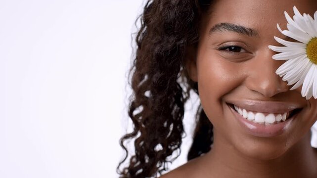 A woman with dark skin and curly hair smiles widely while holding a daisy flower near her right eye. Bright lighting highlights her joyful expression. The background is simple and light.