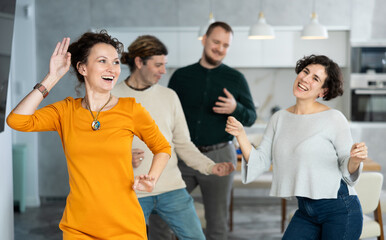 Fototapeta premium Four adult friends dancing and singing at home. Cheerful women and men enjoy music together. Happy party people celebrating friendship