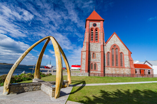 Blue whale mandibles frame Anglican Christ Church Cathedral in Stanley, Falkland Islands, under a clear sky with boats in the background