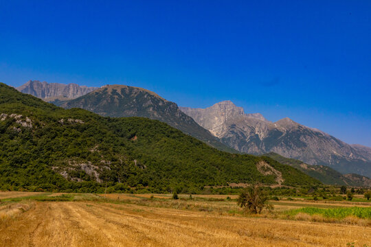 Harvested fields at the base of hills with mountains in the background under a clear sky in southern Albania