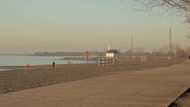 Beach boardwalk with distant walkers in morning haze, lifeguard station and buoys visible along Lake Ontario shoreline, Kew Beach, Toronto, Canada.