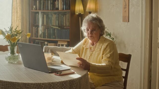 Senior woman with gray hair sitting at table with laptop and smartphone reading billing statement while managing expenses in sunlit living room
