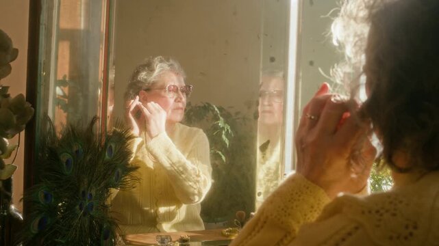 Over shoulder shot of elderly woman with grey hair trying on vintage earrings looking at reflection in mirror while sitting at vanity table in sunlit bedroom