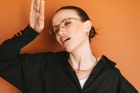 Close up of dramatic young stylish woman with glasses and slicked hair overreacting to stress or bad news, holding hand to forehead with exaggerated suffering face and closed eyes, orange background